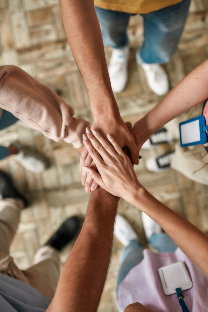 Top view of diverse young volunteers putting their hands on top of each other like a real team while standing in charitable organization office